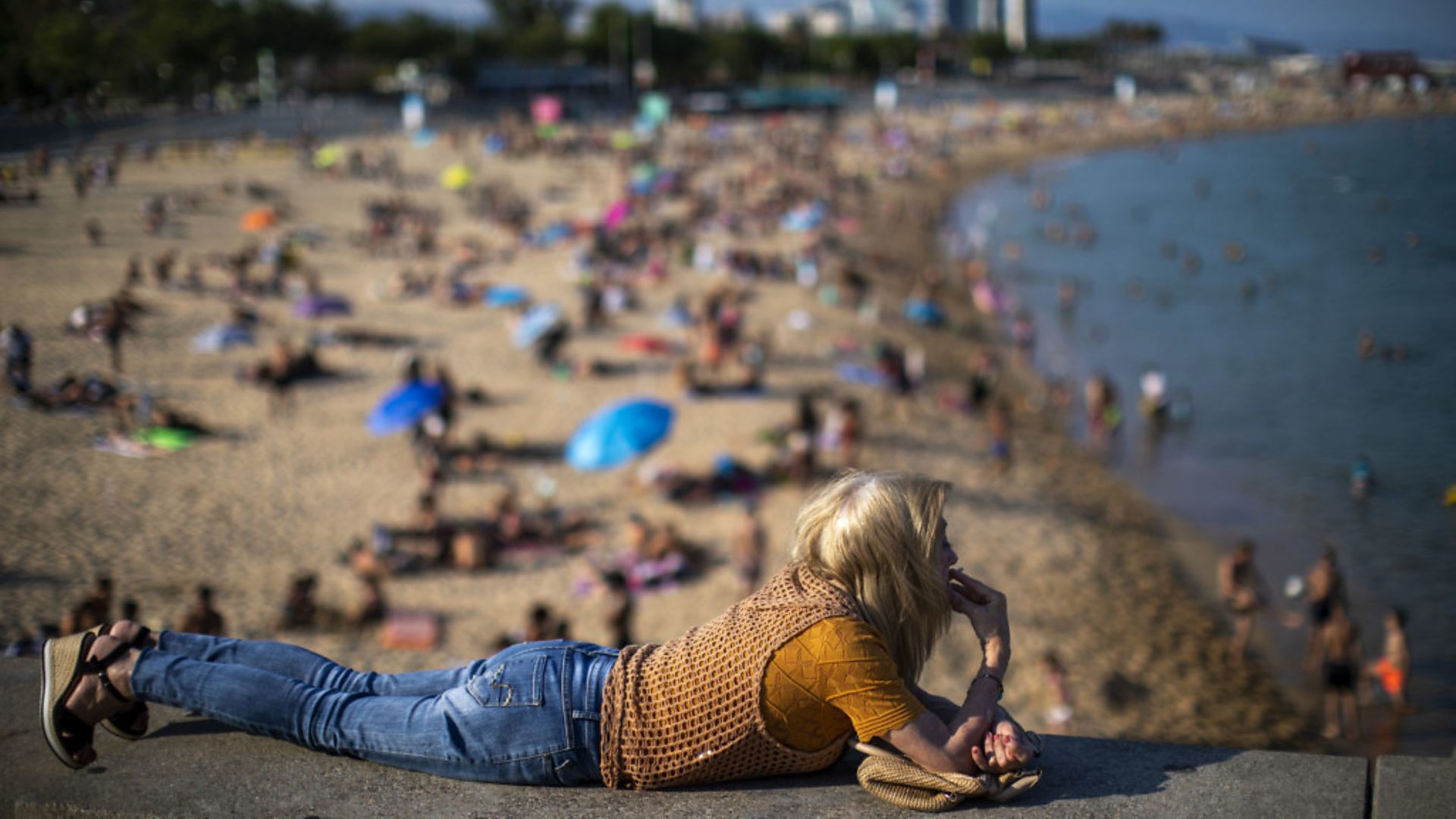 A woman smokes lying on a wall as people enjoy the beach in Barcelona, Spain - Credit: AP